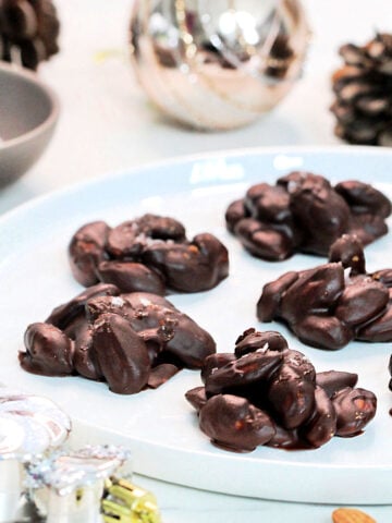 Chocolate almond clusters on a white plate with pinecones and silver ornaments around it.