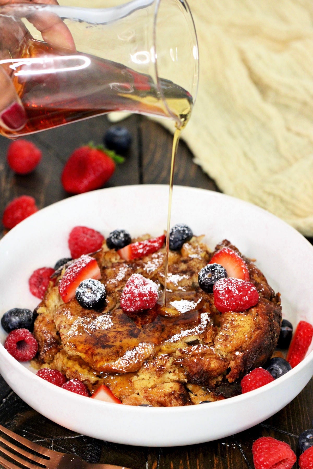 Maple syrup being poured on Crockpot French toast casserole in a white bowl with fresh berries on top.