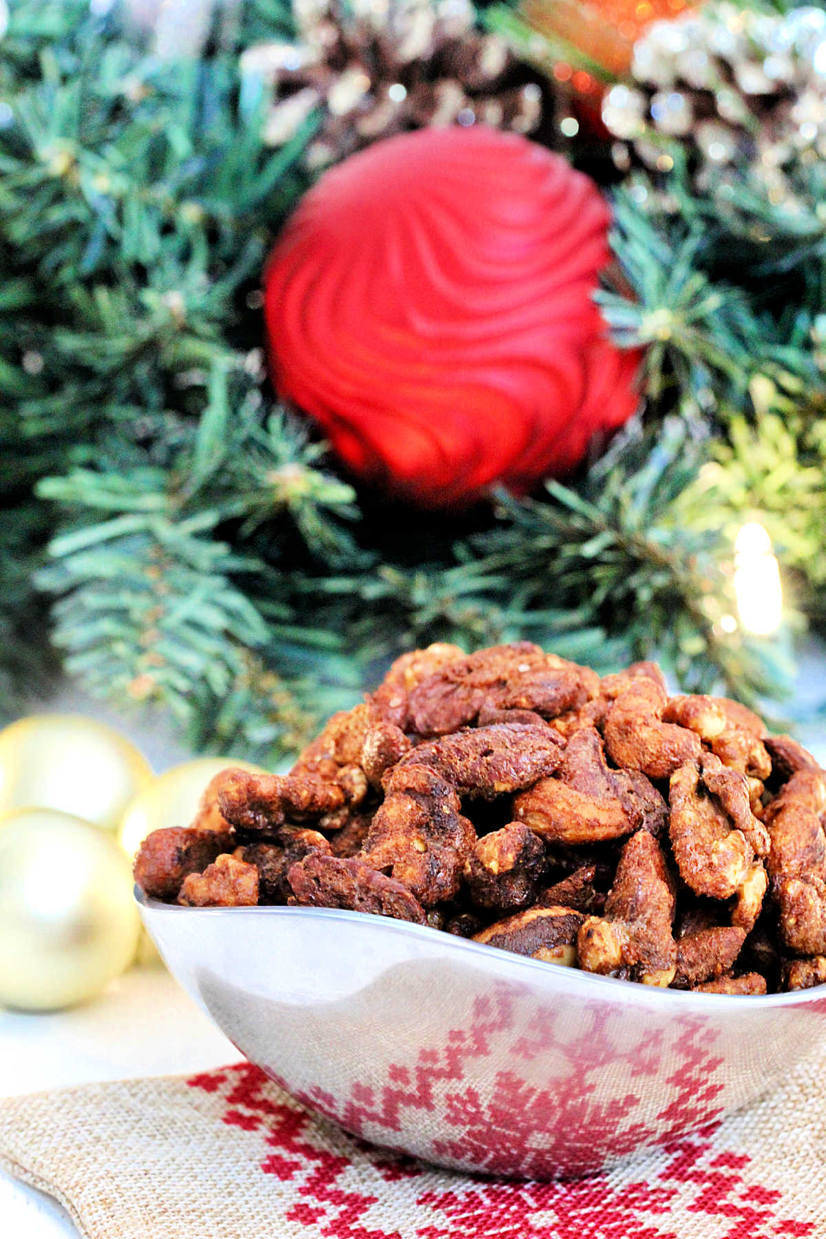 Holiday spiced nuts in a silver bowl in front of a wreath with ornaments.