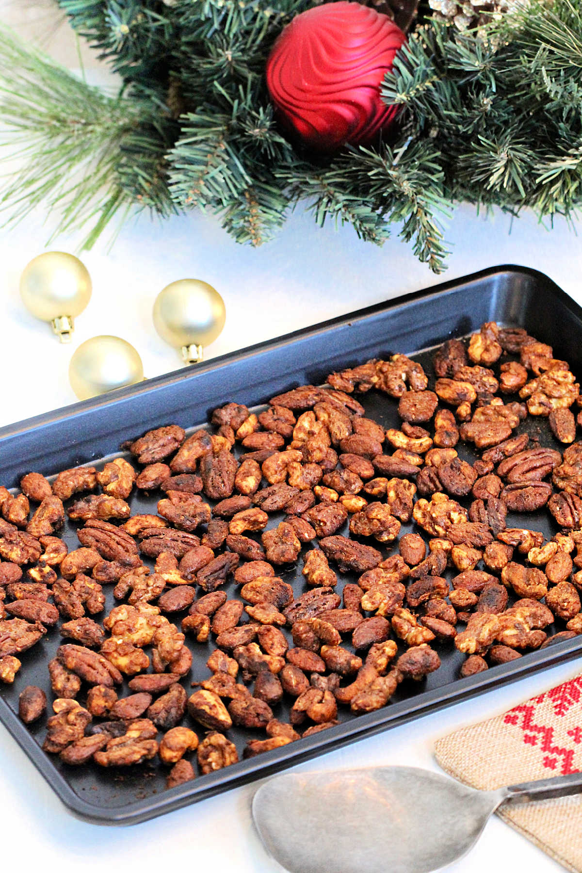 Holiday spiced nuts a black baking sheet in front of a wreath with ornaments around it.