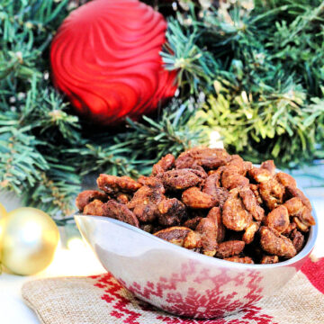 Holiday spiced nuts in a silver bowl in front of a wreath with ornaments.