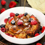 Maple syrup being poured on Crockpot French toast casserole in a white bowl with fresh berries on top.