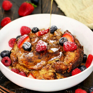 Maple syrup being poured on Crockpot French toast casserole in a white bowl with fresh berries on top.