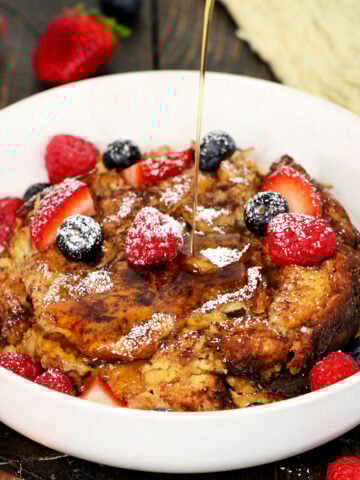 Maple syrup being poured on Crockpot French toast casserole in a white bowl with fresh berries on top.