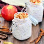 Two jars of apple cinnamon overnight oats.surrounded by an apple, cinnamon sticks, brown napkins and wooden measuring spoons.