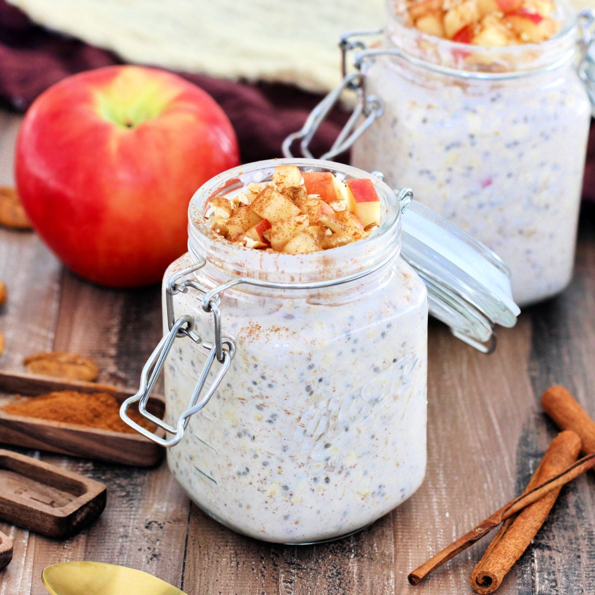 Two jars of apple cinnamon overnight oats.surrounded by an apple, cinnamon sticks, brown napkins and wooden measuring spoons.