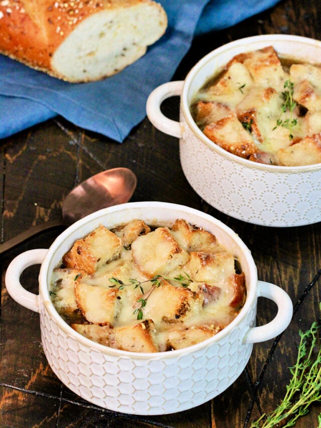 Two bowls of healthy French onion soup on a wooden board with bread, thyme ,a blue napkin and a bronze spoon nearby.