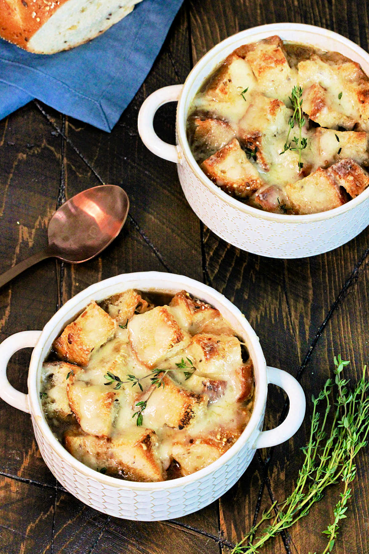 Two bowls of healthy French onion soup on a wooden board with bread, thyme ,a blue napkin and a bronze spoon nearby.