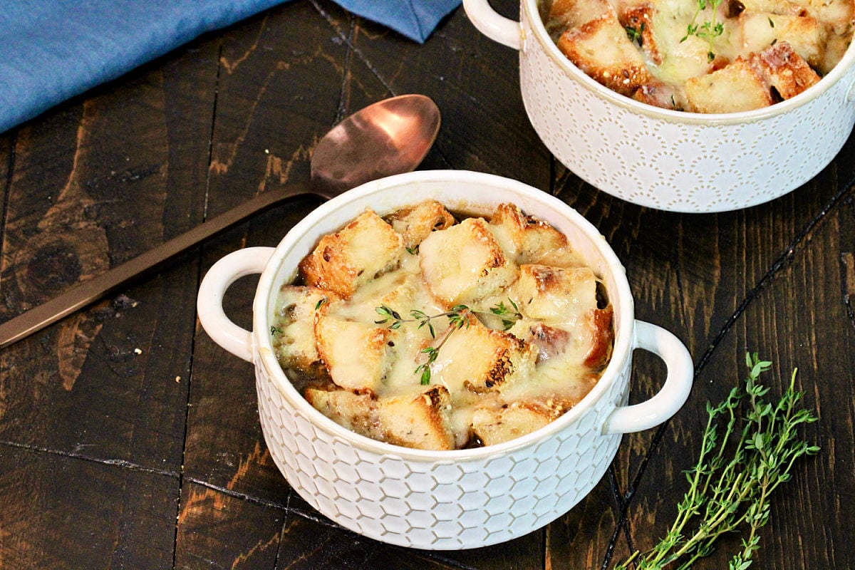Two bowls of healthy French onion soup on a wooden board with thyme ,a blue napkin and a bronze spoon nearby.