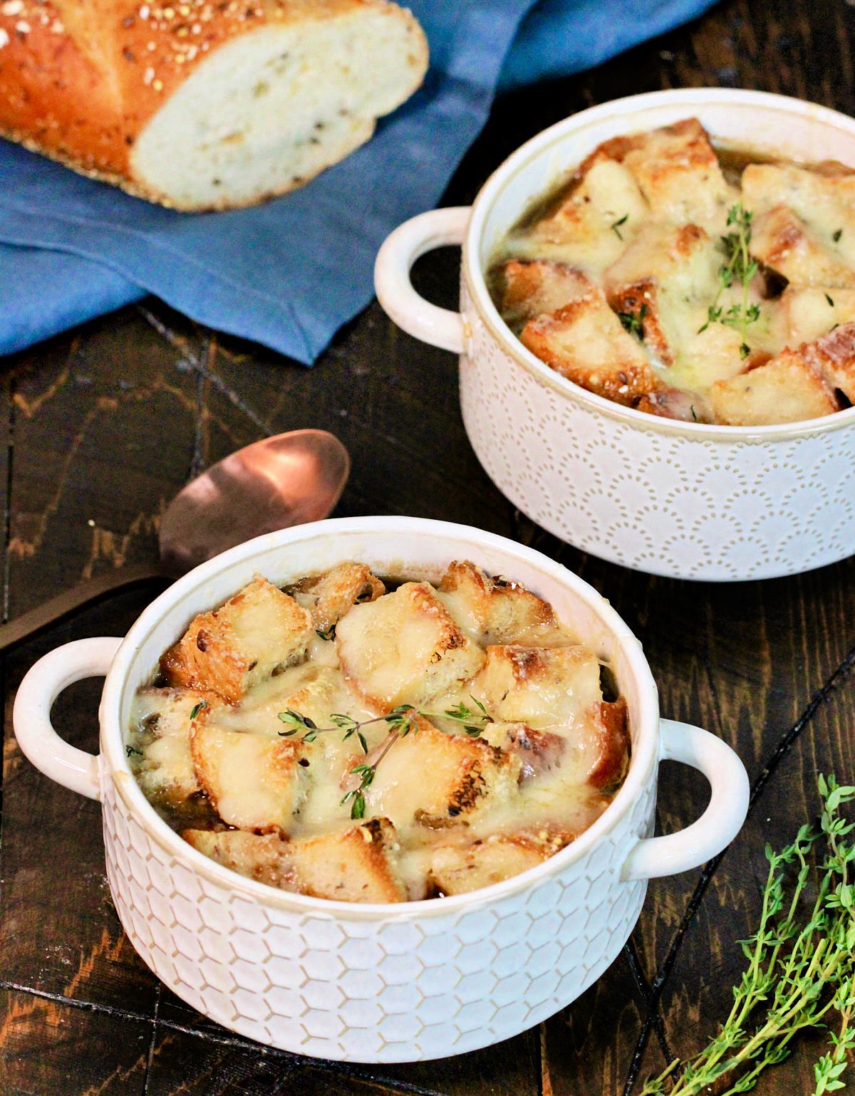 Two bowls of healthy French onion soup on a wooden board with bread, thyme ,a blue napkin and a bronze spoon nearby.