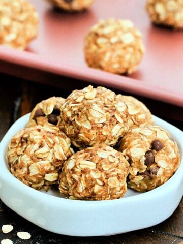 A stack of 3-Ingredient Peanut Butter Oatmeal Balls on a white dish in front of more balls on a pink baking sheet.