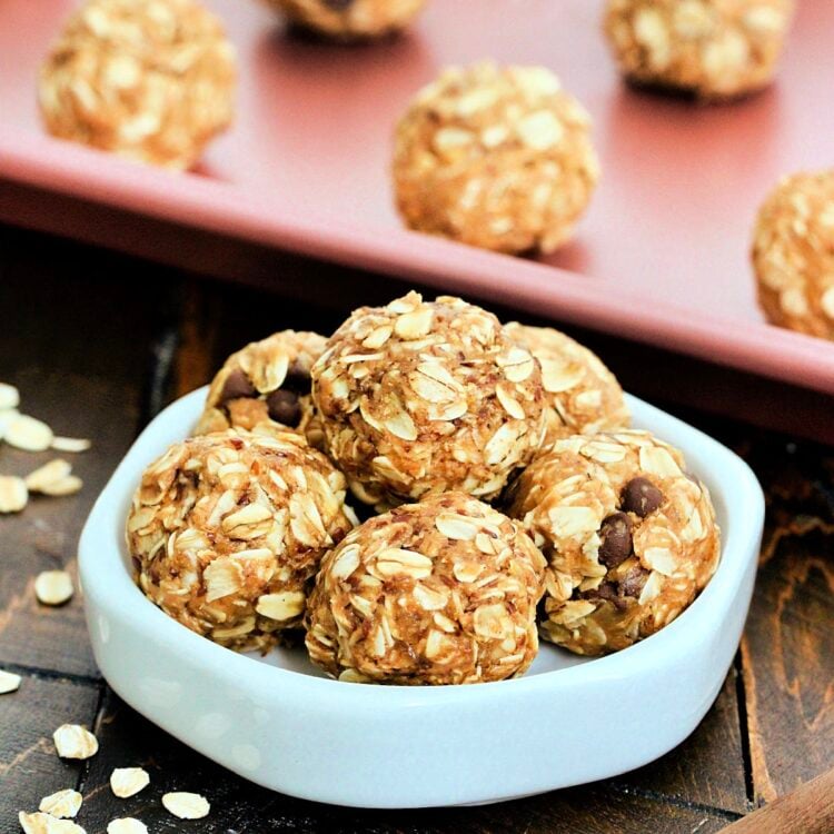 A stack of 3-Ingredient Peanut Butter Oatmeal Balls on a white dish in front of more balls on a pink baking sheet.