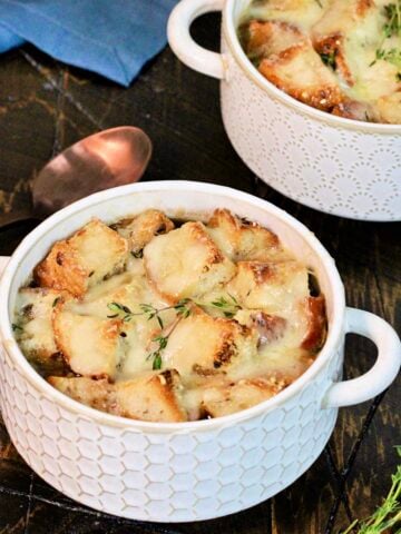 Two bowls of healthy French onion soup on a wooden board with thyme ,a blue napkin and a bronze spoon nearby.