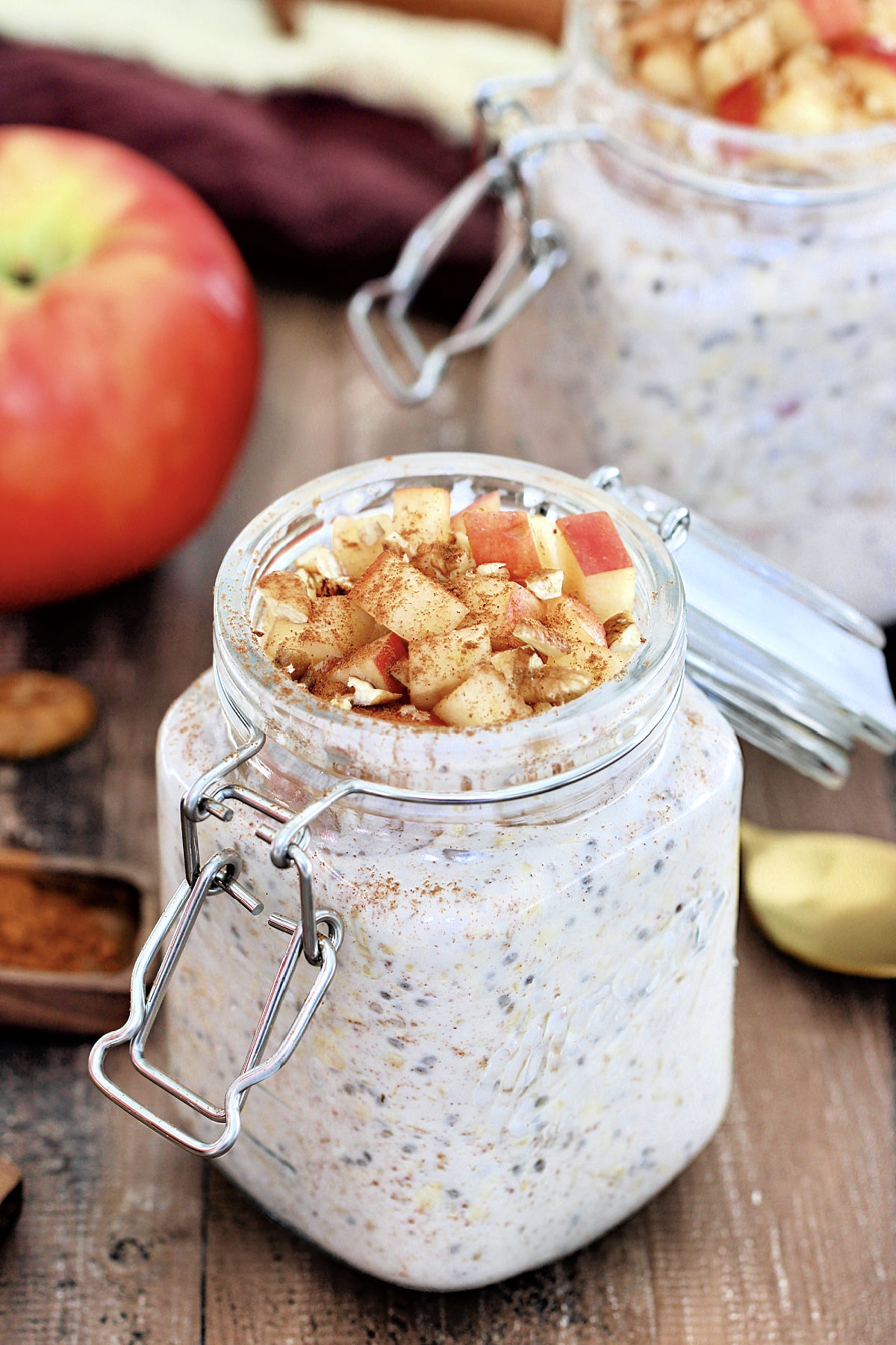 Two jars of apple cinnamon overnight oats.surrounded by an apple, brown napkins and wooden measuring spoons.