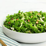 A copycat recipe of Chick-fil-A kale crunch salad in a white bowl with a blue striped napkin and gold fork around it.