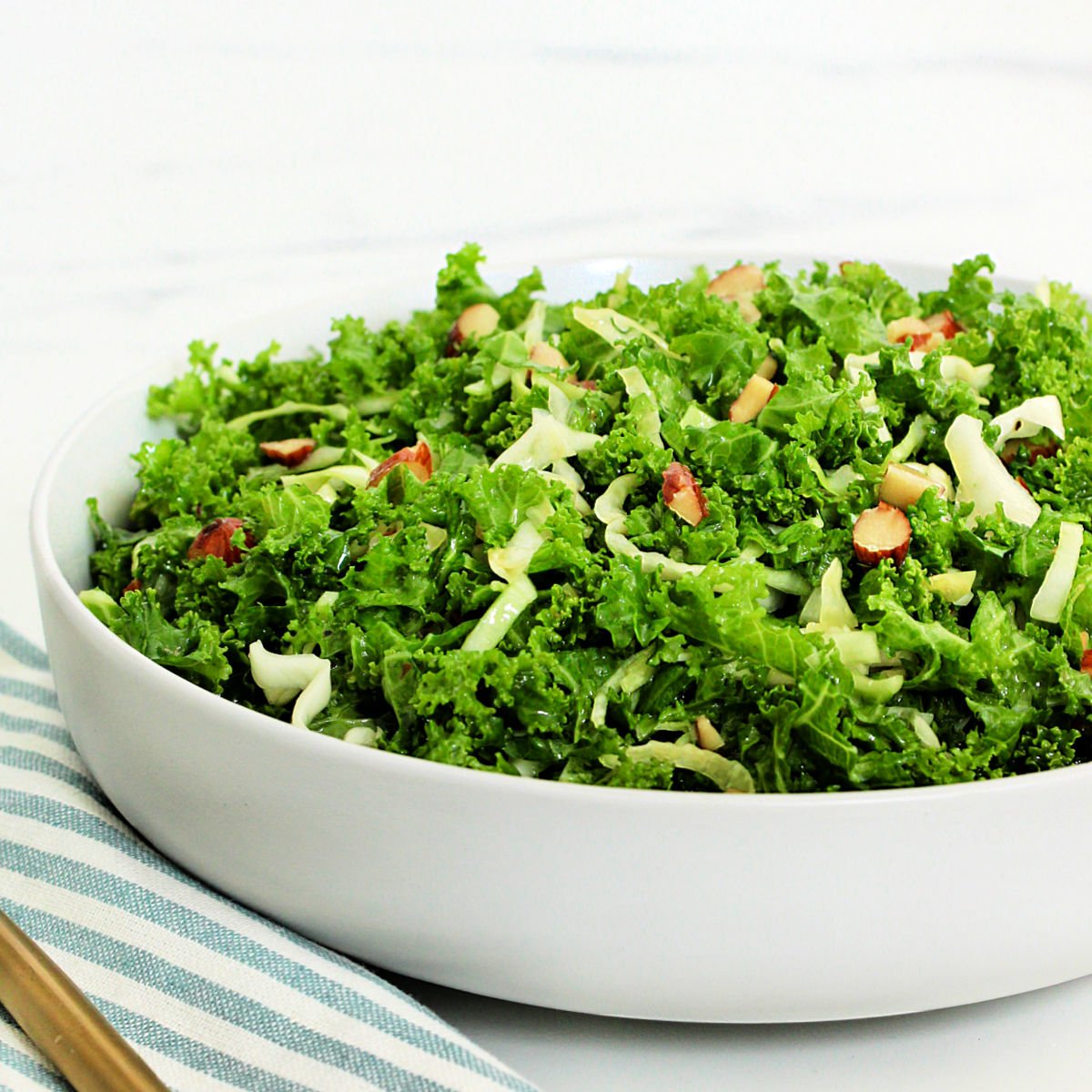A copycat recipe of Chick-fil-A kale crunch salad in a white bowl with a blue striped napkin and gold fork around it.