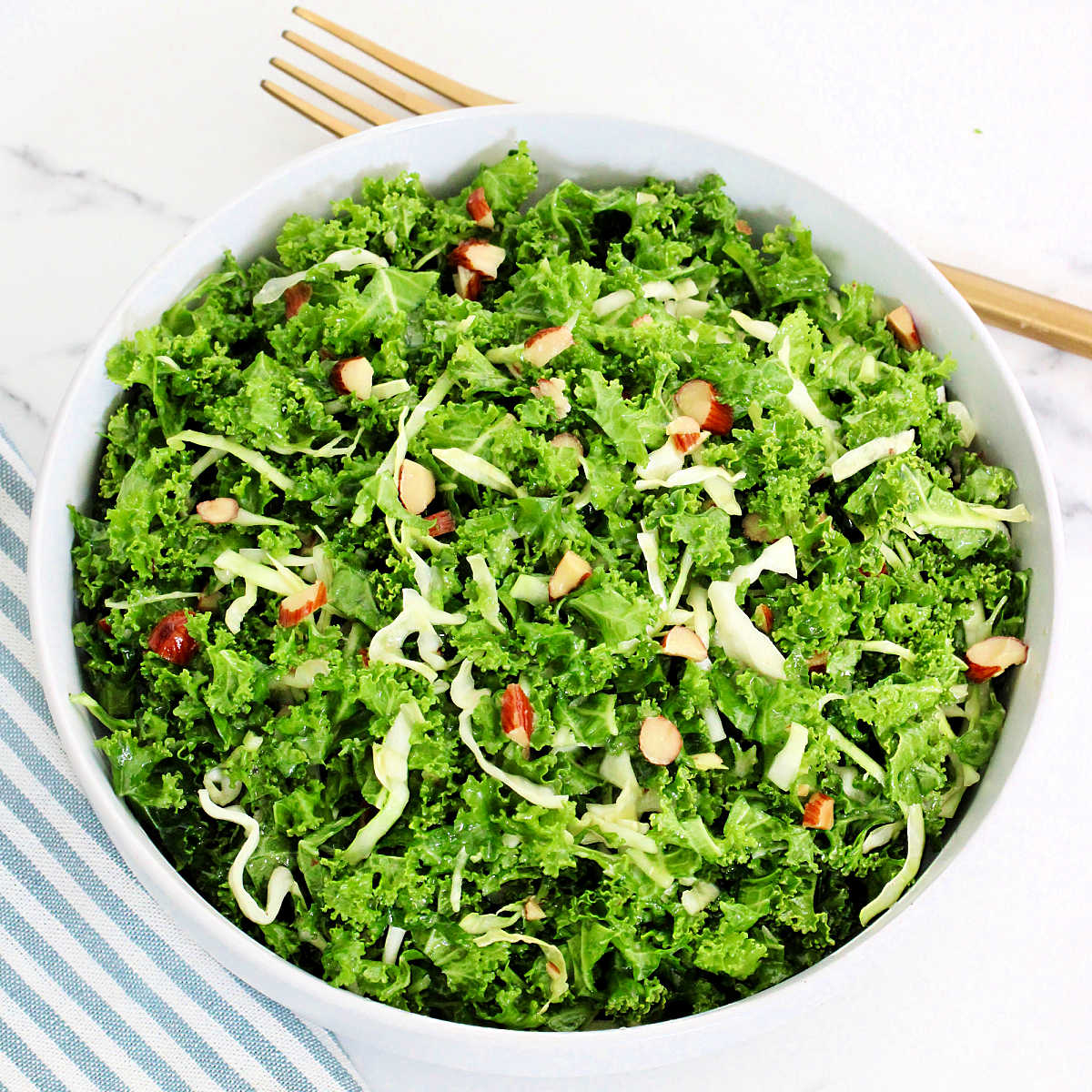 A copycat recipe of Chick-fil-A kale crunch salad in a white bowl with a blue striped napkin and gold fork around it.