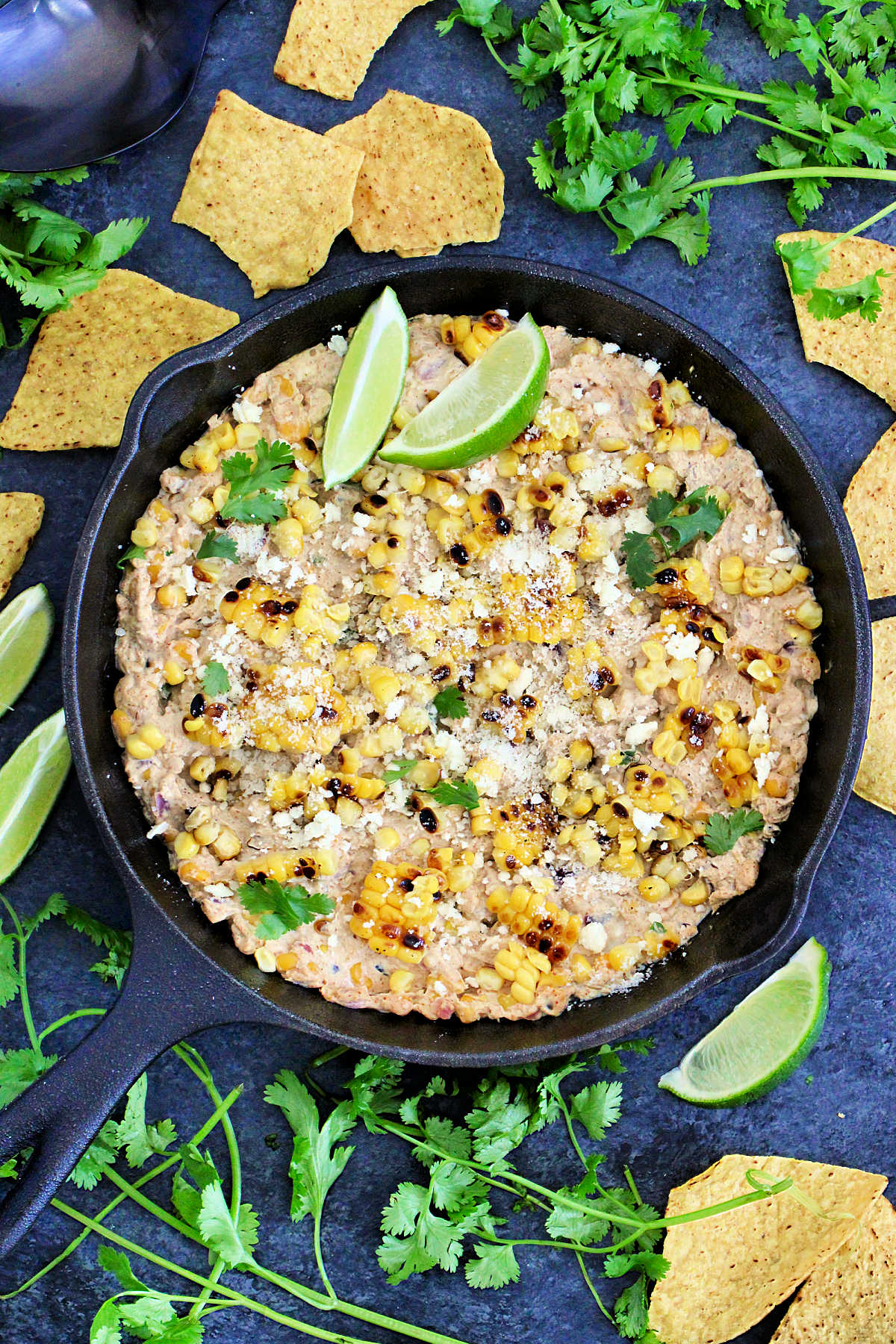 Mexican street corn dip in a cast iron skillet with tortilla chips, lime wedges and cilantro around it.