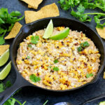 Mexican street corn dip in a cast iron skillet with tortilla chips, lime wedges and cilantro around it.