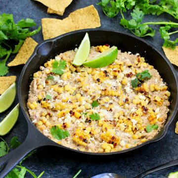 Mexican street corn dip in a cast iron skillet with tortilla chips, lime wedges and cilantro around it.