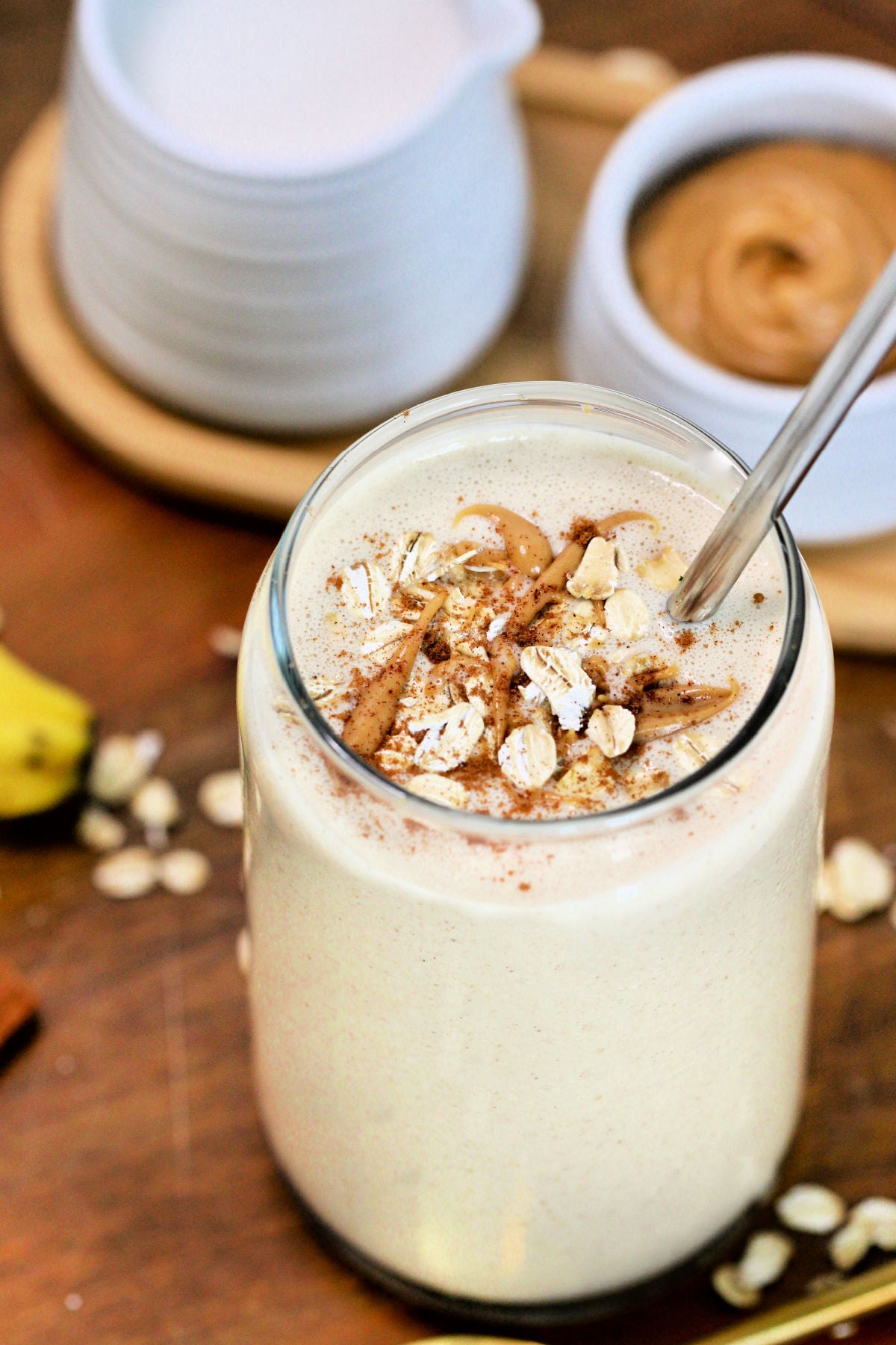 An oatmeal smoothie in a tall glass on a wooden serving platter along with peanut butter, almond milk and a golden spoon.