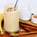 An oatmeal smoothie in a tall glass on a wooden serving platter along with a banana, cinnamon sticks, peanut butter, almond milk and a golden spoon.
