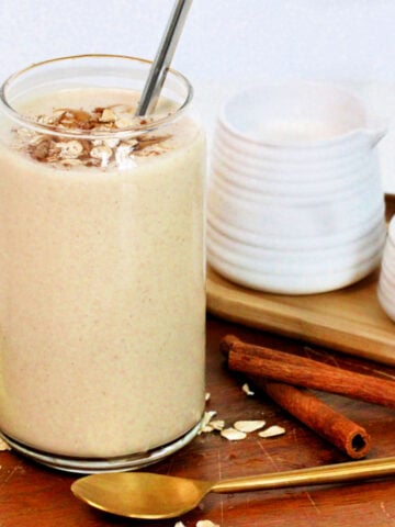 An oatmeal smoothie in a tall glass on a wooden serving platter along with a banana, cinnamon sticks, peanut butter, almond milk and a golden spoon.