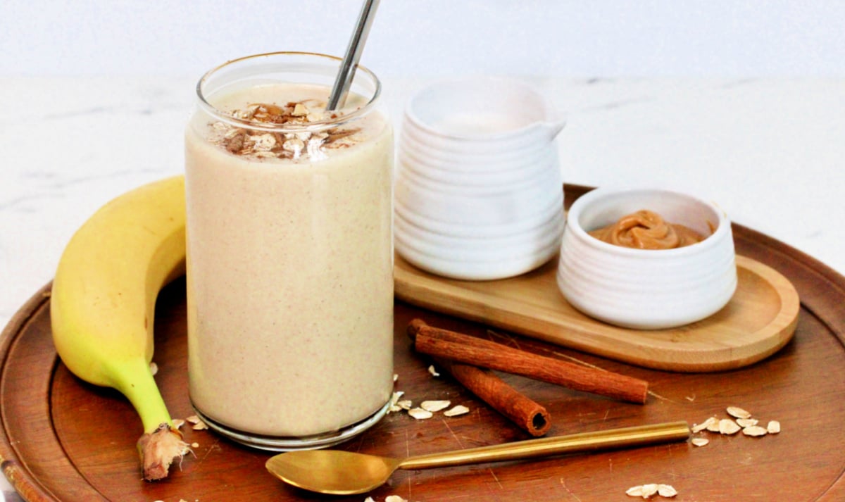 An oatmeal smoothie in a tall glass on a wooden serving platter along with a banana, cinnamon sticks, peanut butter, almond milk and a golden spoon.
