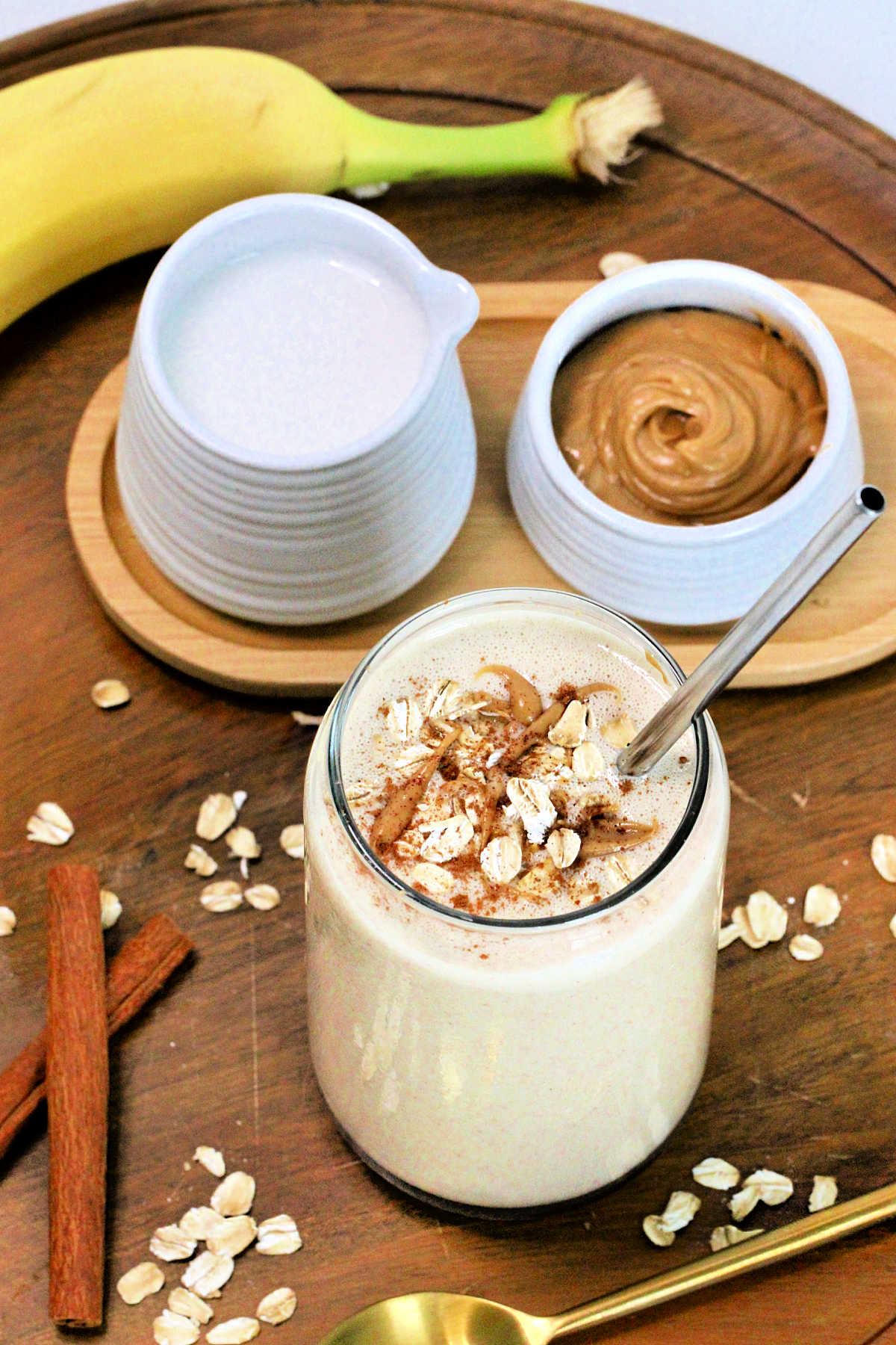 An oatmeal smoothie in a tall glass on a wooden serving platter along with a banana, cinnamon sticks, peanut butter, almond milk and a golden spoon.