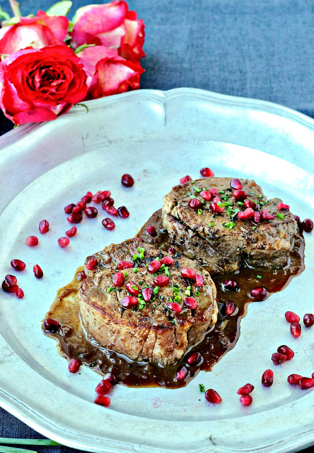Two filet mignons with pomegranate sauce on a silver platter with roses lying nearby.