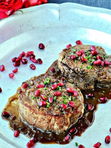 Two filet mignons with pomegranate sauce on a silver platter with roses lying nearby.