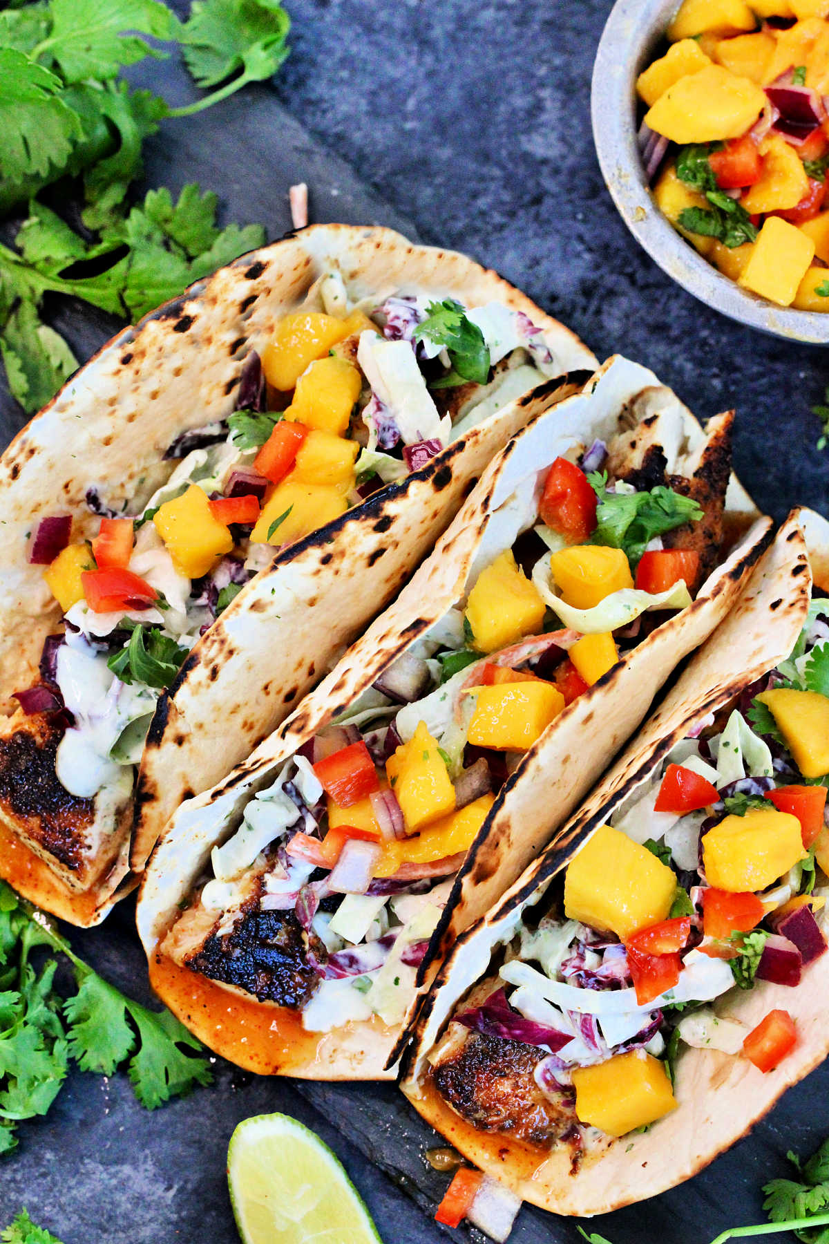 Three mahi mahi tacos surrounded by cilantro and a bowl of mango salsa.