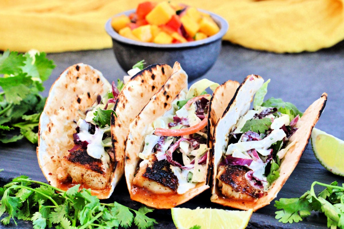 Three mahi mahi tacos surrounded by cilantro and a bowl of mango salsa.