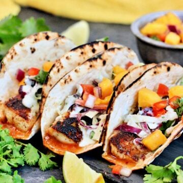 Three mahi mahi tacos surrounded by cilantro and a bowl of mango salsa.