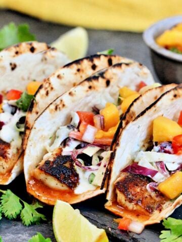Three mahi mahi tacos surrounded by cilantro and a bowl of mango salsa.