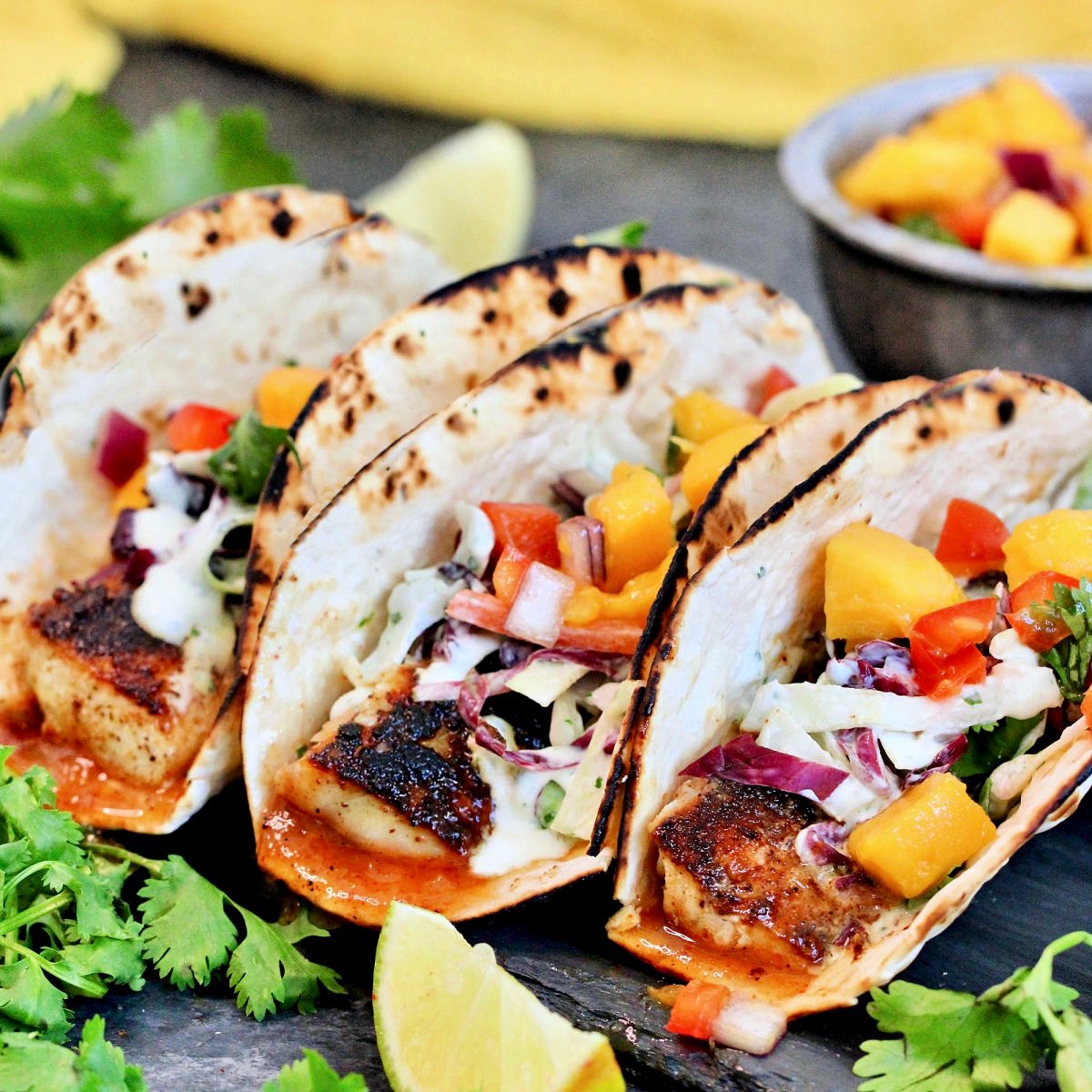 Three mahi mahi tacos surrounded by cilantro and a bowl of mango salsa.