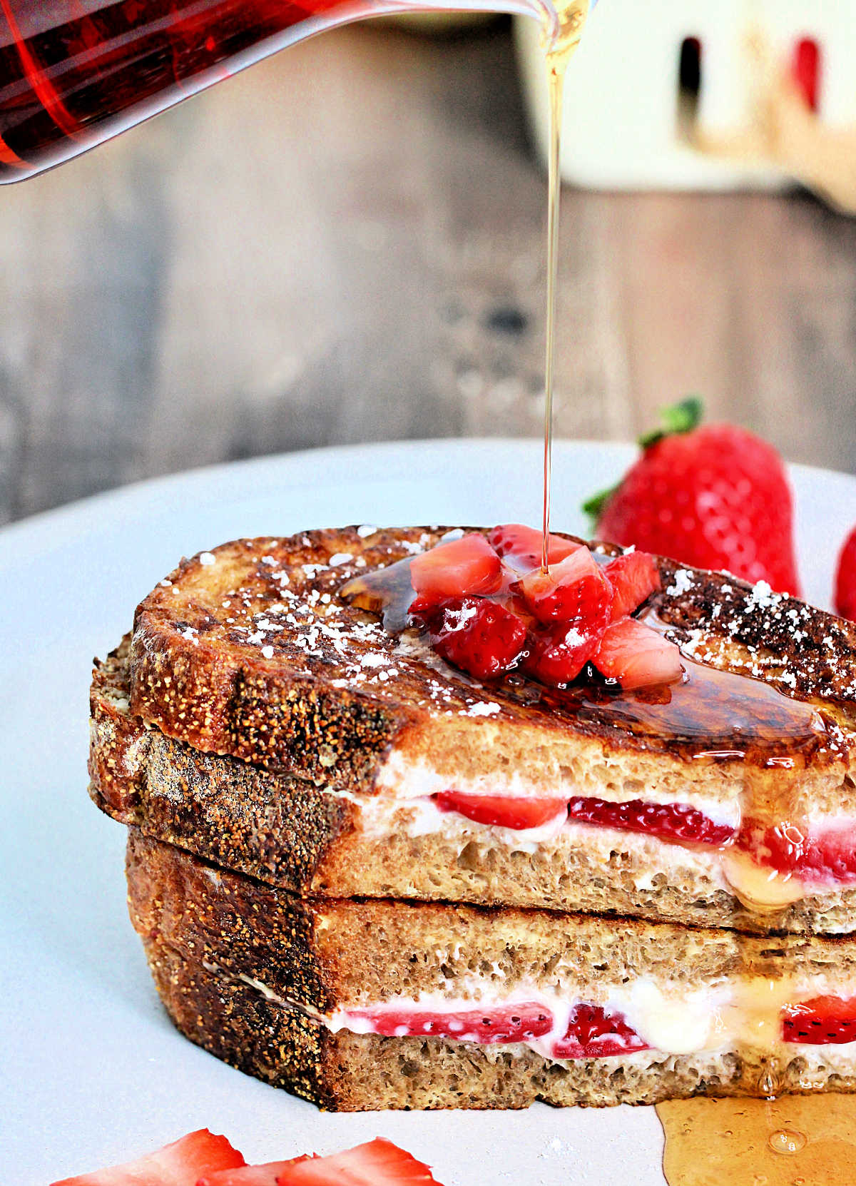 Maple syrup being poured onto two pieces of strawberry stuffed French toast stacked on a white plate.