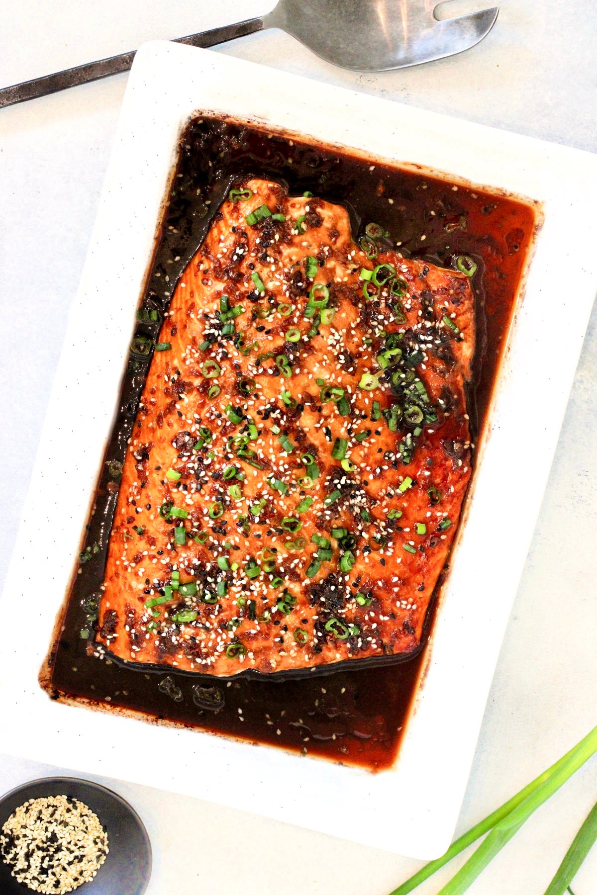 Honey ginger salmon in a white serving platter with sesame seeds and a silver serving utensil around it.