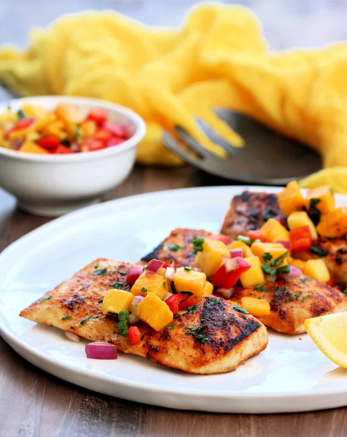 Grilled mahi mahi topped with mango salsa on a white plate with a bowl of mango salsa and a yellow napkin in the background.