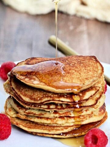 Maple syrup being poured on a stack of 3-Ingredient banana protein pancakes on a white plate with raspberries and a gold folk around them.