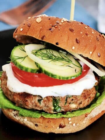 Greek turkey burger on a black plate with a blue napkin and a fork in the background.
