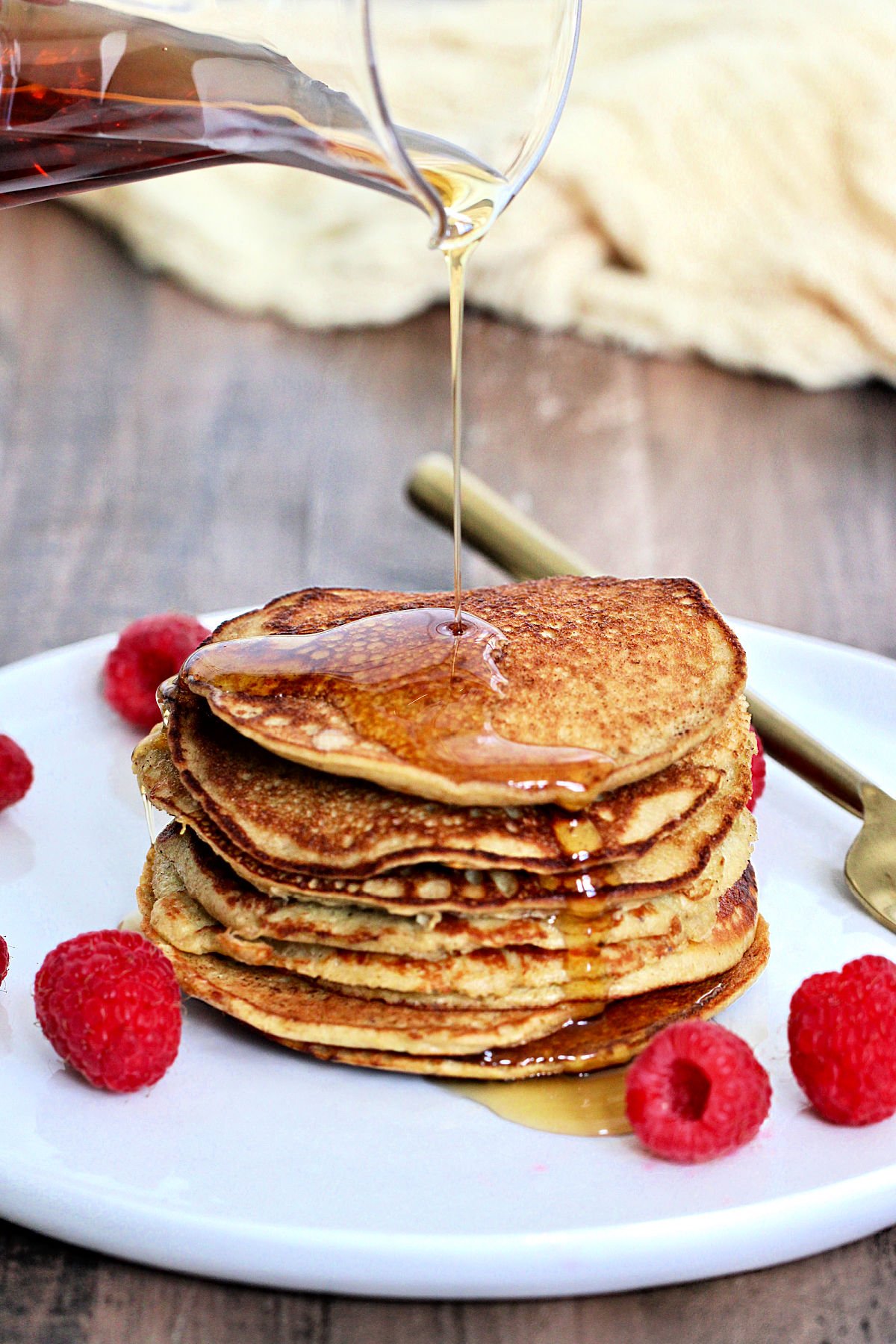 Maple syrup being poured on a stack of 3-Ingredient banana protein pancakes on a white plate with raspberries and a gold folk around them.