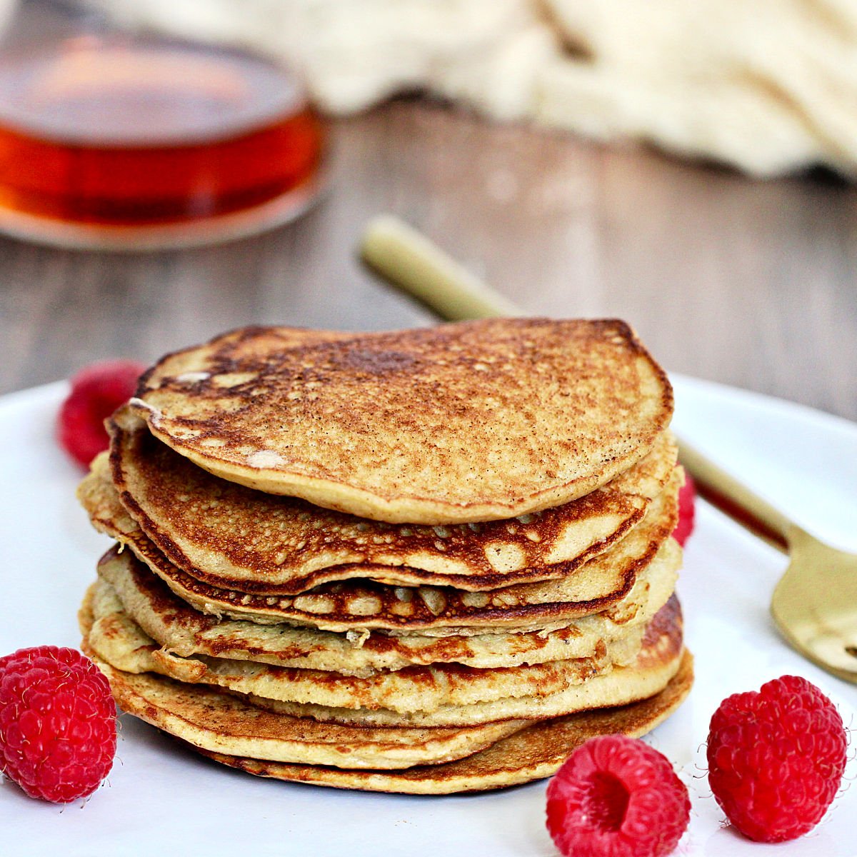 A stack of 3-Ingredient banana protein pancakes on a white plate with raspberries and a gold folk around them.