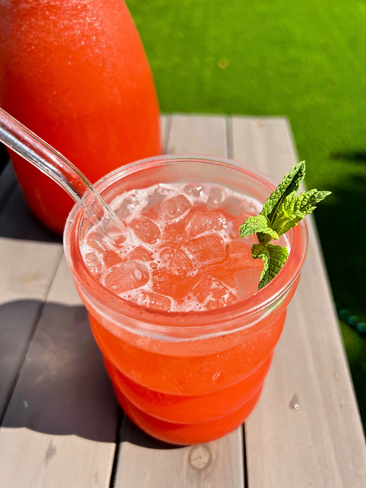 A glass of strawberry pineapple lemonade on an outdoor table with pitcher of lemonade behind it.