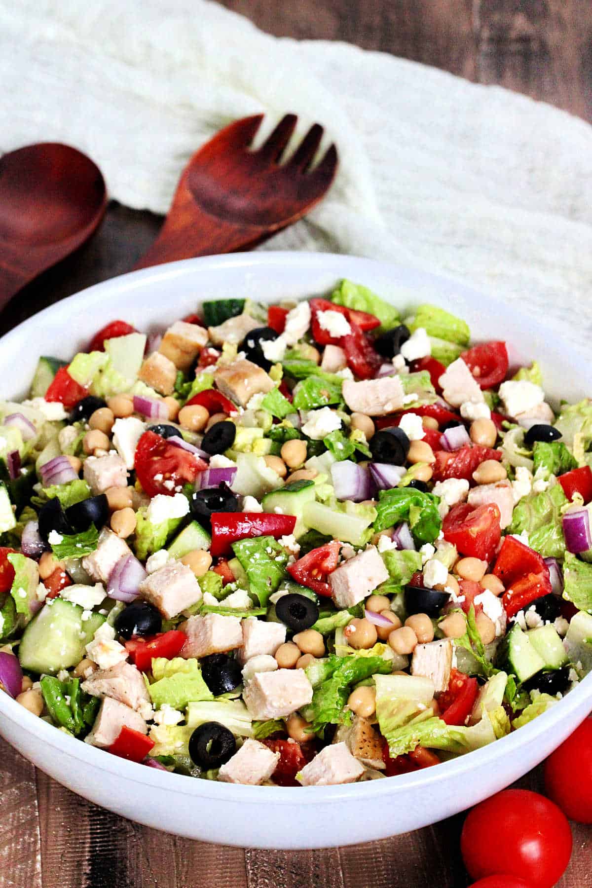 Mediterranean chopped salad in a white bowl with tomoatoes and woodern utensils nearby.