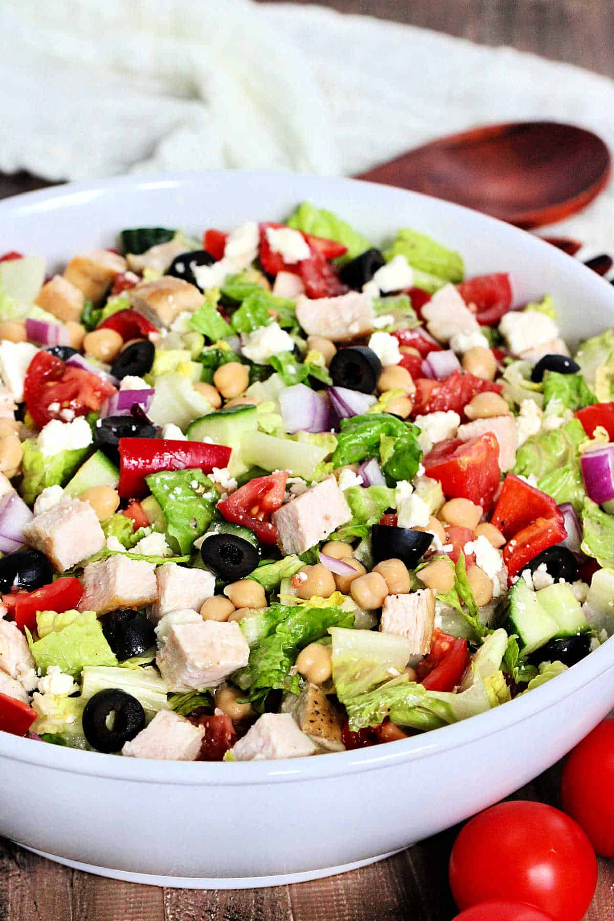 Mediterranean chopped salad in a white bowl with tomoatoes and woodern utensils nearby.