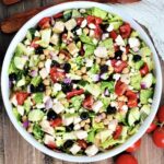 Mediterranean chopped salad in a white bowl with tomoatoes and woodern utensils nearby.