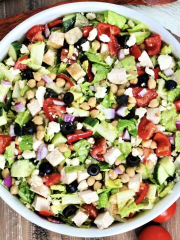 Mediterranean chopped salad in a white bowl with tomoatoes and woodern utensils nearby.