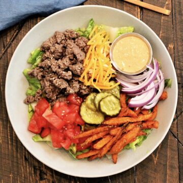 A burger bowl on a wooden board with a blue napkin and bronze fork nearby.