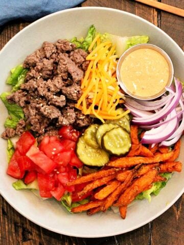 A burger bowl on a wooden board with a blue napkin and bronze fork nearby.